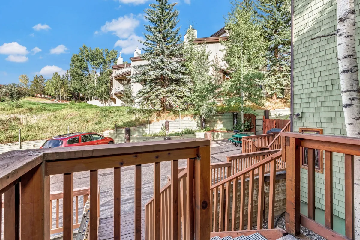 A peaceful wooden deck looking out onto green trees, with a red car parked nearby and surrounding buildings visible. The deck is ideal for relaxing outdoors or catching up with a neighbor, and the visible parking supports the mention of on-site parking.
