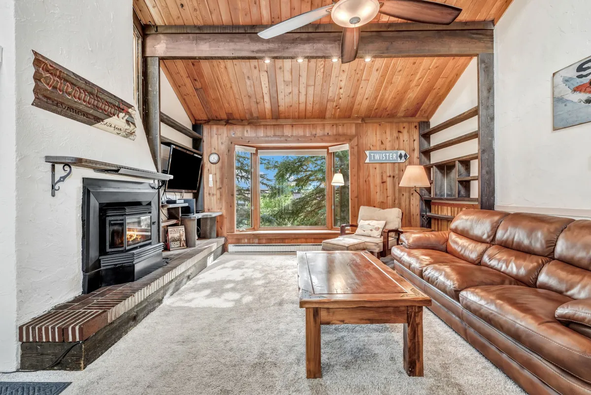 This image features a plush leather sofa positioned in front of a wood-burning fireplace, with a large window that lets in abundant natural light. Exposed wooden beams and a coffee table add to the rustic mountain ambiance, and the overall scene suggests a welcoming, communal atmosphere for relaxing, reading, or gathering with family and friends.