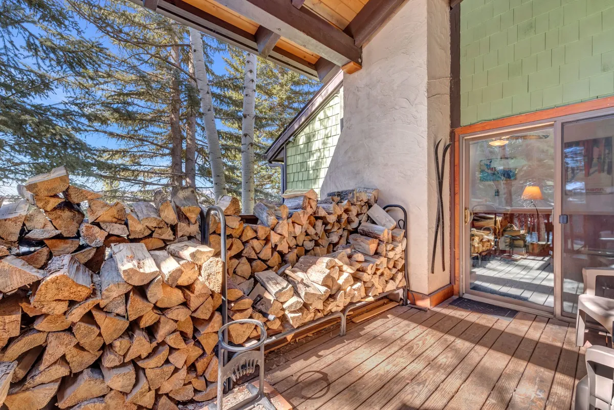 A rustic exterior scene featuring a wooden deck with a stack of firewood, a sliding glass door leading inside, and trees surrounding the space—creating an inviting transition between indoor comfort and outdoor play.