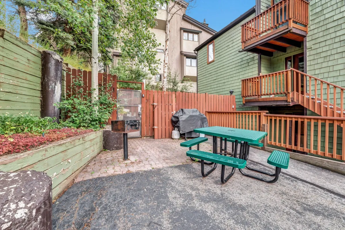 A charming outdoor area featuring a picnic table and a barbecue grill set beside a lush garden. The space is designed for casual outdoor dining and socializing, surrounded by greenery that enhances privacy and comfort.