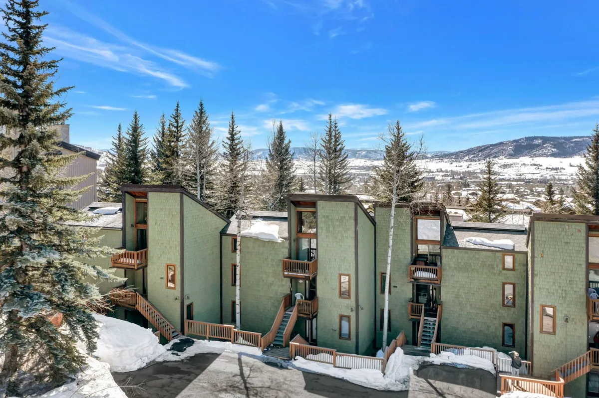 A tranquil exterior view of the multi-level property nestled among pine trees, with fresh snow blanketing the ground and balconies inviting relaxation after a day outside. The clear blue sky and visible ski tracks evoke the excitement of being close to ski activities.