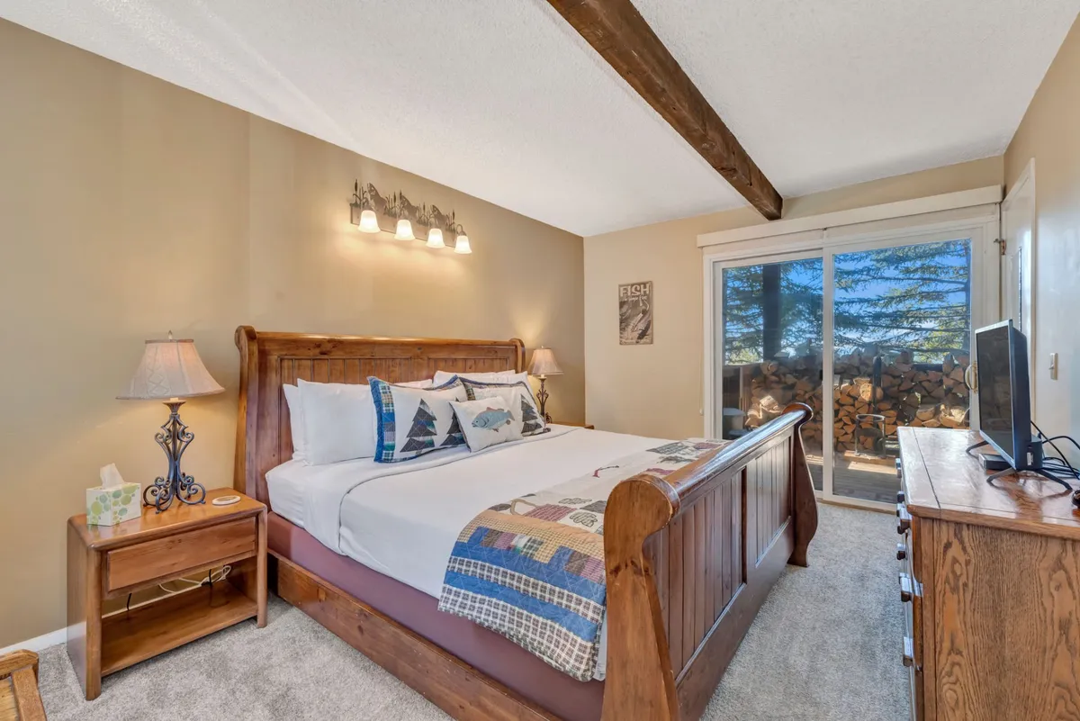 A warmly lit king bedroom with a sleigh bed topped with decorative pillows, a nightstand and lamp, and a sliding door that opens to a view of a neatly stacked woodpile. The cozy ambiance is enhanced by soft lighting and natural wood finishes.