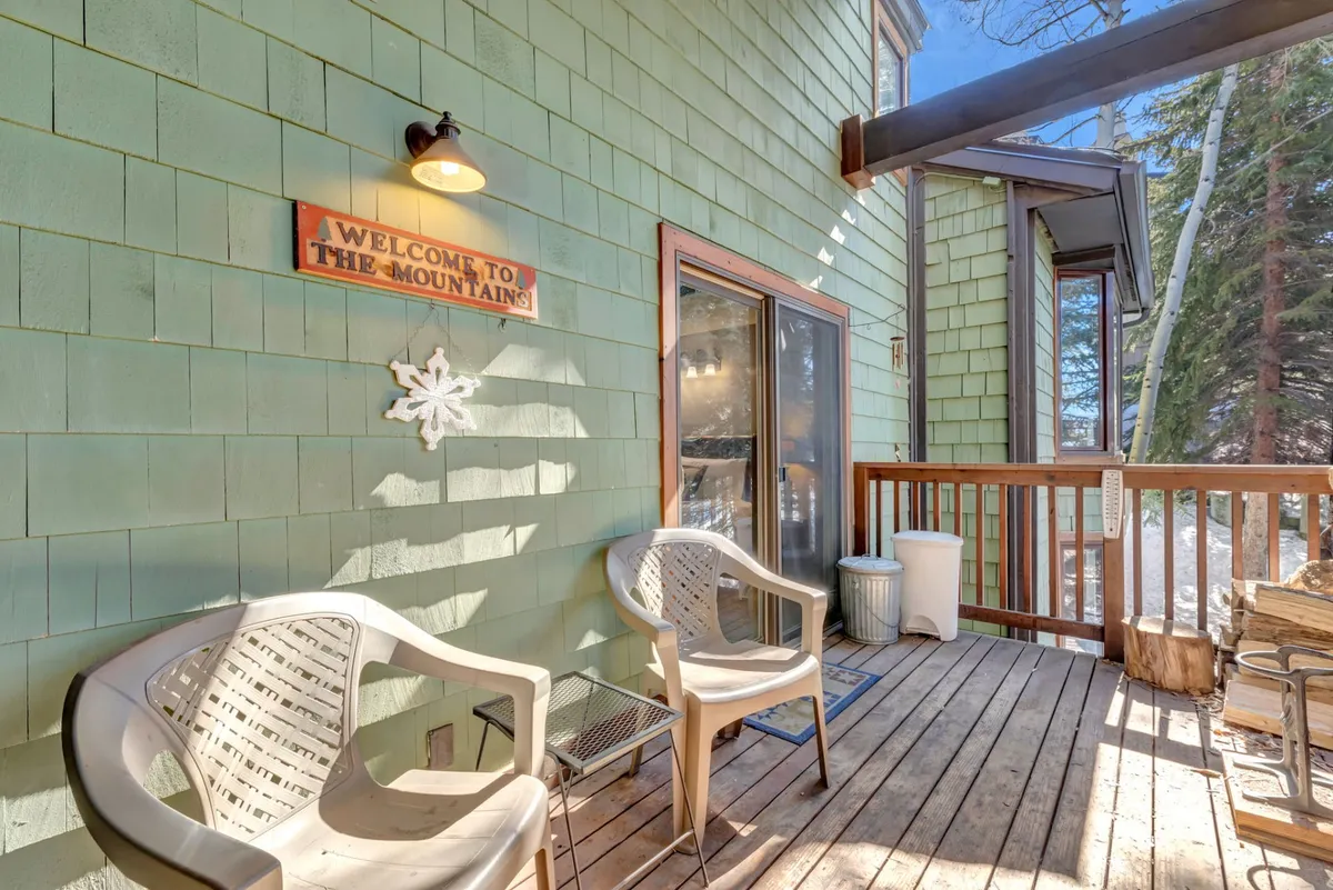 A welcoming entrance area with a 'Welcome' sign, wooden chairs, railing, snow on the ground, and pine trees in the background. The inviting setting feels friendly and intimate, with plenty of space for socializing or enjoying a peaceful moment outdoors.