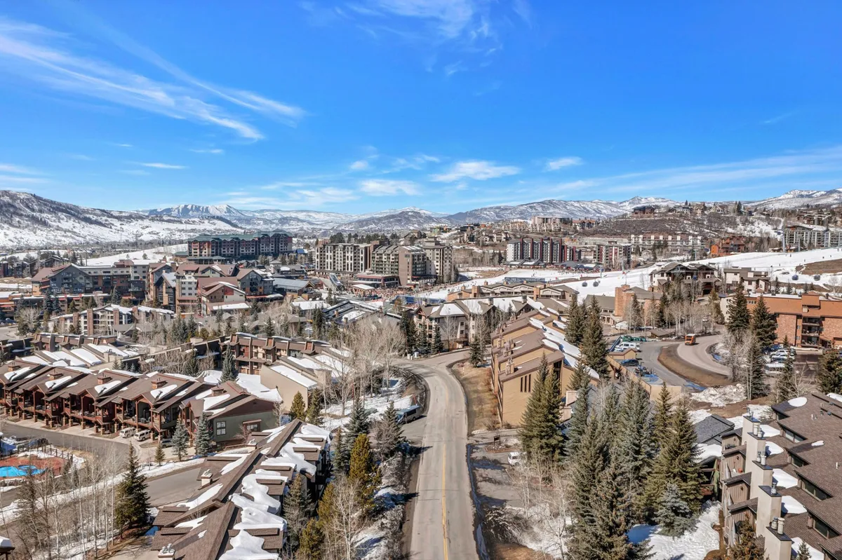 A picturesque winter scene featuring snow-covered rooftops, distant mountains, a winding road, and rows of cozy townhouses nestled among trees—conveying a tranquil and inviting neighborhood atmosphere.