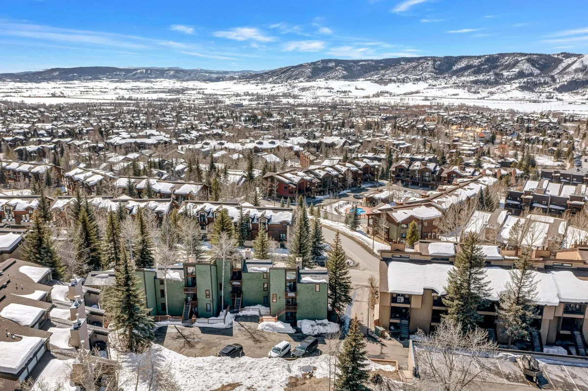 A picturesque exterior view featuring snow-covered rooftops and green architectural accents, set against a backdrop of surrounding mountains. The scene evokes a sense of arrival and hints at the winter recreation that defines Steamboat Springs.