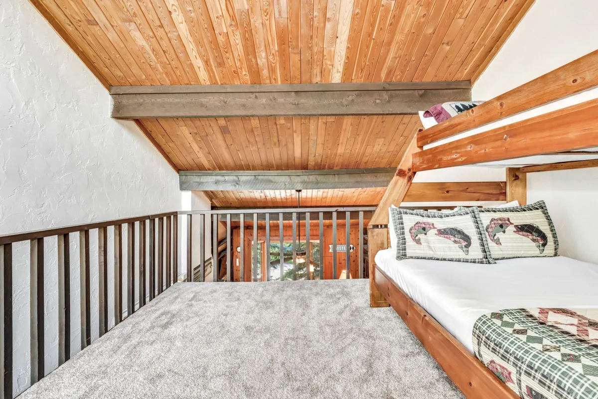 A cozy open loft furnished with a twin-over-full bunk bed, plush carpet, wooden ceiling, and colorful decorative pillows. The space feels playful and welcoming, ideal for children or extra guests.