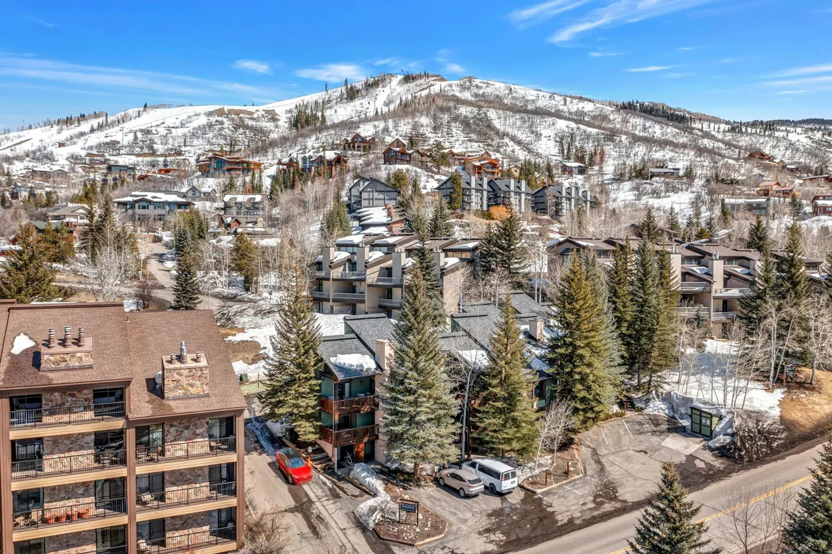 A picturesque scene featuring snow-capped mountains rising in the background, framed by tall pine trees and the modern architecture of nearby residential buildings. The snow-covered landscape captures the spirit of winter adventure and proximity to the slopes.