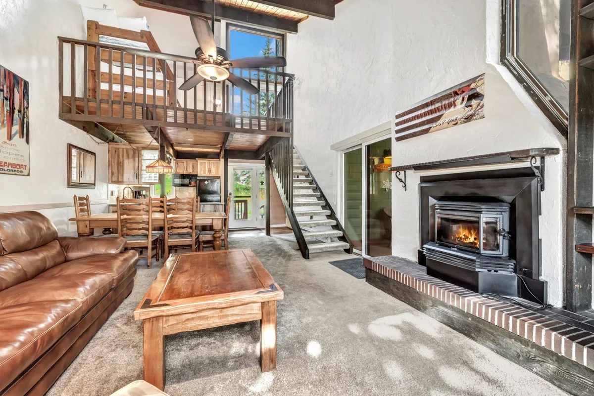 This interior shot captures a seamless connection between the living room and dining area. The space includes a leather sofa, a wooden dining table, and a fireplace against the back wall. The image also features a staircase, contributing to the sense of open-concept design and communal gathering.