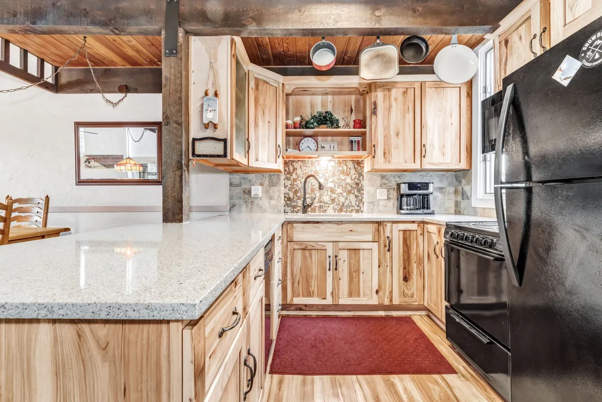 A cozy kitchen scene featuring a granite countertop, wooden cabinetry, black appliances, a decorative shelf with plants, and a red accent rug. The space feels rustic and inviting—ideal for both everyday meals and special gatherings.