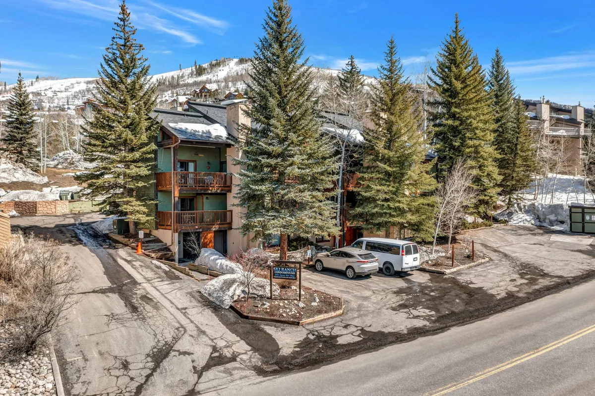 The image showcases the exterior of a mountain condo nestled among tall pine trees, with the building's rustic-chic architecture standing out against a backdrop of stunning mountain scenery. The tranquil setting is complemented by a spacious parking area, inviting guests to arrive and settle in easily. The overall mood is peaceful, with hints of cozy outdoor relaxation.